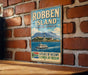 A vintage-style metal sign with the text "ROBBEN ISLAND SOUTH AFRICA" and an illustration of a boat on a body of water, mounted on a brick wall.