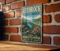 A vintage-style metal sign for Rothrock State Forest in Pennsylvania, featuring a scenic landscape with pine trees, a river, and pink flowers.