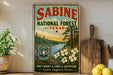 A vintage-style metal sign for Sabine National Forest in Texas, featuring a scenic landscape with pine trees, a river, and flowers.