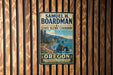 A vintage-style metal sign for the State Scenic Corridor in Oregon, featuring a scenic coastal landscape with mountains, cliffs, and a winding path along the water.