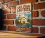 A vintage-style metal sign for San Juan Bautista State Historic Park, featuring an illustration of a historic building and surrounded by lush greenery.