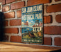 A vintage-style metal sign for San Juan Island National Historical Park, featuring an illustration of the park's historic buildings and a ship, mounted on a brick wall.