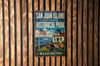 A vintage-style metal sign for San Juan Island National Historical Park, featuring an American flag, a lighthouse, and a ship, mounted on a wooden wall.