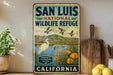 A vintage-style metal sign for San Luis National Wildlife Refuge in California, featuring an illustration of a bird, flowers, and mountains.