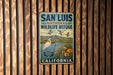 A vintage metal sign for the San Luis National Wildlife Refuge in California, featuring a bird and flowers, mounted on a wooden wall.