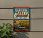 A vintage-style sign for San Luis Valley, Colorado, featuring a scenic landscape with mountains, a river, and a small building.