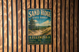 A vintage metal sign for Sand Ridge State Forest in Illinois, featuring a scenic landscape with a pine tree and a dirt path.
