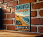 A vintage-style metal sign with the text "SANDWICH HARBOUR" and "NAMIBIA" hangs on a brick wall.
