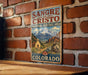 A vintage-style metal sign for the Sangre de Cristo National Heritage Area in Colorado, featuring a mountain landscape and a church.