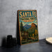 A vintage metal sign for Santa Fe National Forest in New Mexico, featuring a scenic mountain landscape and text promoting the forest's natural beauty and open spaces.