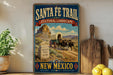 A vintage-style poster advertising the Santa Fe Trail in New Mexico, featuring a wagon train traveling through a desert landscape with mountains in the background.