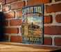 A vintage-style metal sign for the Santa Fe Trail in New Mexico, featuring a wagon traveling through a desert landscape with mountains in the background.