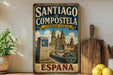 A vintage-style sign for "SANTIAGO de COMPOSTELA CIUDAD VIEJA" (Santiago Compostela, Cienada Vieja) is displayed on a wall, with a potted plant and kitchen utensils visible in the foreground.