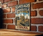 A vintage metal sign for "SANTIAGO de COMPOSTELA" in Spain, featuring an illustration of the city's landmarks and a clock.