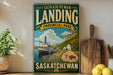 A vintage-style sign for "Saskatchewan Landing" in the Canadian province of Saskatchewan, featuring a scenic image of a boat on a lake and a small village in the background.