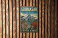 a vintage metal sign with a mountain landscape and a river, mounted on a wooden wall.