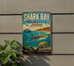 A vintage-style sign for Shark Bay, Western Australia, featuring a shark, a boat, and other marine life, mounted on a wall.