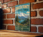 A vintage-style metal sign for Shasta-Trinity National Forest in California, featuring a mountain landscape with pine trees and a lake.