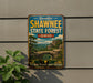 A vintage-style metal sign advertising Shawnee State Forest in Ohio, featuring a scenic landscape with a bridge and trees.