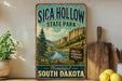 A vintage-style sign for Sica Hollow State Park in South Dakota, featuring a scenic mountain landscape with a river and pine trees.