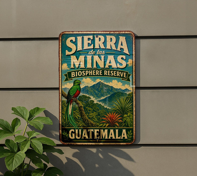 A vintage-style metal sign with the text "SIERRA de las MINAS BIOSPHERE RESERVE GUATEMALA" and an illustration of a colorful bird perched on a tree branch, set against a backdrop of lush tropical vegetation.