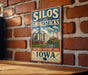 A vintage-style metal sign with the text "SILOS and SMOKE stacks" and "NATIONAL HERITAGE AREA IOWA" is displayed on a brick wall.