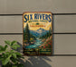 a vintage-style metal sign with a scenic illustration of a river surrounded by mountains and pine trees, mounted on a wall.