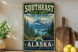 A vintage-style sign for Southeast State Forest in Alaska, featuring a scenic mountain landscape with pine trees and a lake.