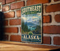 A vintage-style metal sign with a mountain landscape and pine trees, mounted on a brick wall.