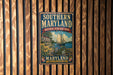 A vintage metal sign for the Southern Maryland National Heritage Area, featuring a sailboat and a house, is displayed on a wooden wall.