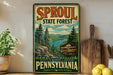 a vintage-style sign for the Pennsylvania State Forest, with a scenic illustration of a forest and a river. The sign is displayed on a shelf next to a wooden cutting board and some potted plants.