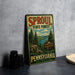A vintage-style metal sign for the Pennsylvania State Forest, featuring a scenic illustration of pine trees and a river, is displayed on a gray countertop.