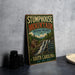 A vintage metal sign for Stumphouse Mountain Heritage Preserve, featuring a scenic illustration of a waterfall and a wooden cabin, is displayed on a gray countertop next to a stack of black camera lenses.