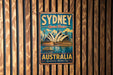 A vintage metal sign with the text "SYDNEY Opera House" and "AUSTRALIA Spectacular Harbour City" is displayed on a wooden wall.