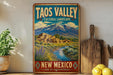 A vintage-style sign for the Taos Valley Cultural Landscape in New Mexico, featuring a scenic landscape with mountains, adobe buildings, and a river.