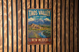 A vintage-style metal sign for the Taos Valley in New Mexico, featuring a mountain landscape and a small town.