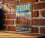 A vintage-style metal sign for Teide National Park in Spain, featuring a mountain landscape with a prominent peak and a blue sky with clouds.