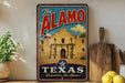 A vintage-style metal sign with the text "The Alamo" and "Texas Remember the Alamo!" displayed prominently, featuring an illustration of the historic Alamo building.