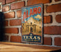 A vintage-style metal sign with the text "The Alamo" and "Texas Remember the Alamo!" displayed on a brick wall.