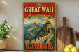 A vintage-style metal sign with the text "The Great Wall of China" and a picture of the iconic wall, placed on a shelf next to a wooden cutting board and some potted plants.