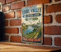 A vintage metal sign with text and a painting of a river and castle in the background, mounted on a brick wall.