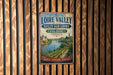 A vintage metal sign with text and a painting of a valley, mounted on a wooden wall.