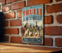 A vintage-style metal sign with the text "The TRULLI of ALBEROBELO ITALY" and an illustration of a village with thatched roofs, hanging on a brick wall.