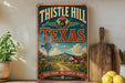 A vintage-style metal sign for Thistle Hill, Texas, featuring a scenic countryside landscape with a windmill, flowers, and a house.