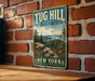 A vintage-style metal sign for Tug Hill State Forest in New York, featuring a scenic landscape with pine trees, a river, and a mountain.