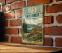 A vintage metal sign for Tuller Hill State Forest in New York, featuring a scenic landscape with pine trees, a lake, and mountains.