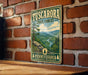 A vintage-style metal sign for Tuscarora State Forest in Pennsylvania, featuring a scenic mountain landscape with pine trees and a winding river.