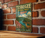 A vintage-style metal sign for Tuskegee National Forest in Alabama, featuring a scenic landscape with a deer and a river.