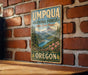 A vintage-style metal sign for the Umpqua National Forest in Oregon, featuring a scenic landscape with mountains, pine trees, and a river.