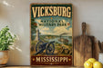 A vintage-style metal sign for Vickburg National Military Park in Mississippi, featuring a cannon, a river, and a steamboat, with the text "VICKSBURG NATIONAL MILITARY PARK" and "MISSISSIPPI" prominently displayed.